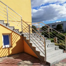 Zewnętrzne schody z szarego granitu z nierdzewną balustradą, prowadzące do wejścia budynku o żółtej elewacji, widoczne w słoneczny dzień na tle błękitnego nieba z chmurami.