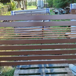 ALU-STAL - Gotowy, brązowy panel ogrodzeniowy z poziomymi szczeblami, leżący na palecie. W tle widoczne inne elementy ogrodzenia, częściowo owinięte folią.