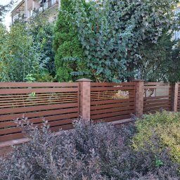 ALU-STAL - Nowoczesne ogrodzenie z poziomych, brązowych paneli i ceglanych słupków, otoczone zielenią. Widok na budynek w tle. Ujęcie z poziomu gruntu.