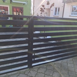 ALU-STAL - Nowoczesna, pozioma brama wjazdowa w kolorze antracytowym, z widocznym mechanizmem zamka. Tło stanowi budynek mieszkalny z zieloną elewacją i elementami dekoracyjnymi.