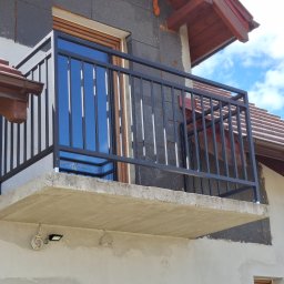 ALU-STAL - Czarna, metalowa balustrada balkonowa z pionowymi szczebelkami, zamontowana na betonowej płycie balkonu. Widok z dołu, fragment elewacji budynku w tle.