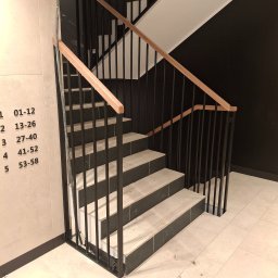ALU-STAL - Metalowe schody z drewnianą poręczą w nowoczesnym budynku. Widoczne numery mieszkań na ścianie. Minimalistyczny design, czarne barierki i jasne stopnie.