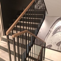 ALU-STAL - Nowoczesne schody z czarną metalową balustradą i drewnianym poręczą, widok z góry. Na ścianie dekoracyjny motyw architektoniczny w odcieniach szarości.