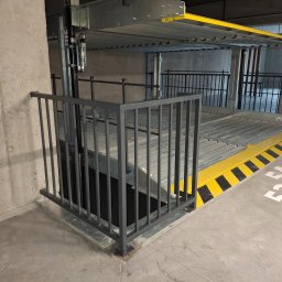 ALU-STAL - Szara, metalowa barierka zabezpieczająca platformę parkingu wielopoziomowego. Żółto-czarne pasy ostrzegawcze na krawędzi platformy. Numeracja miejsc parkingowych na posadzce.
