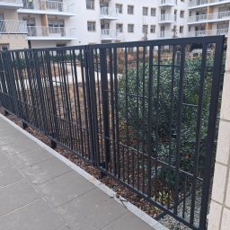 ALU-STAL - Czarne, metalowe ogrodzenie panelowe na tle nowoczesnego budynku mieszkalnego i zieleni. Widok z poziomu chodnika.