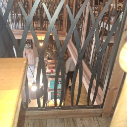 ALU-STAL - Metalowa balustrada o dekoracyjnym wzorze, widok z góry na wnętrze restauracji. Elementy spawane, drewniane wykończenie. Nietypowy kształt prętów.