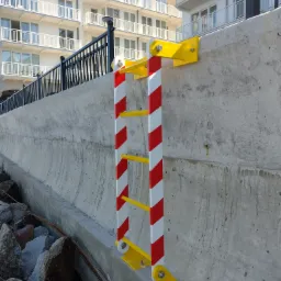 Przymocowana do betonowego nabrzeża drabina z żółtymi szczeblami i biało-czerwonymi pasami na bokach, u dołu kamienie, w tle budynek z balkonami.