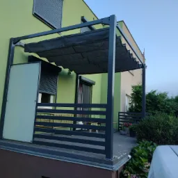 Nowoczesna pergola z szarą, przesuwaną tkaniną zacieniającą, wsparta na metalowej konstrukcji, przylegająca do zielonego budynku z roletami zewnętrznymi i szklaną osłoną boczną.