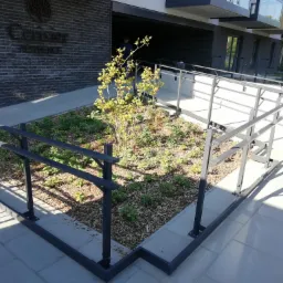 Metalowa balustrada zabezpieczająca rabatę z roślinami przed budynkiem mieszkalnym z rampą dla niepełnosprawnych.