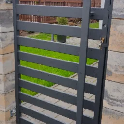 Szara furtka z poziomych profili stalowych, zamontowana między słupkami z bloczków betonowych, widok na trawnik i zabudowania w tle.