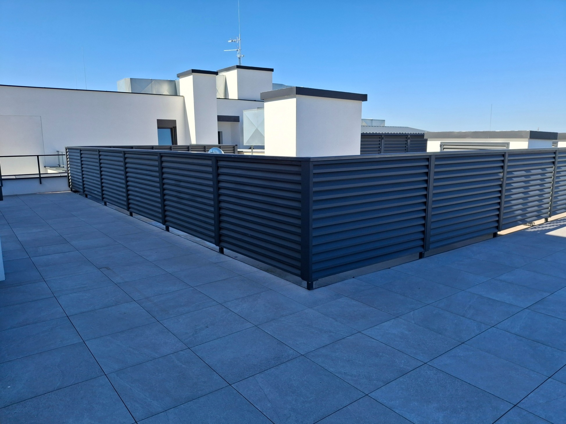 Nowoczesne, poziome, metalowe panele ogrodzeniowe w kolorze grafitowym, zamontowane na tarasie budynku z szarą, kwadratową kostką brukową, na tle białej architektury i błękitnego nieba.