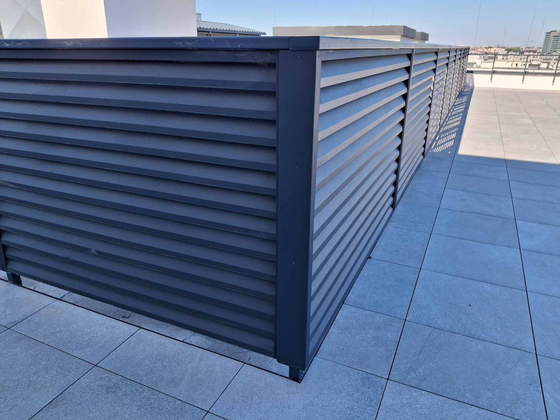 Szara osłona balkonowa z poziomych listew na tle miejskiego krajobrazu, widoczna z perspektywy narożnika balkonu wyłożonego szarymi płytkami.