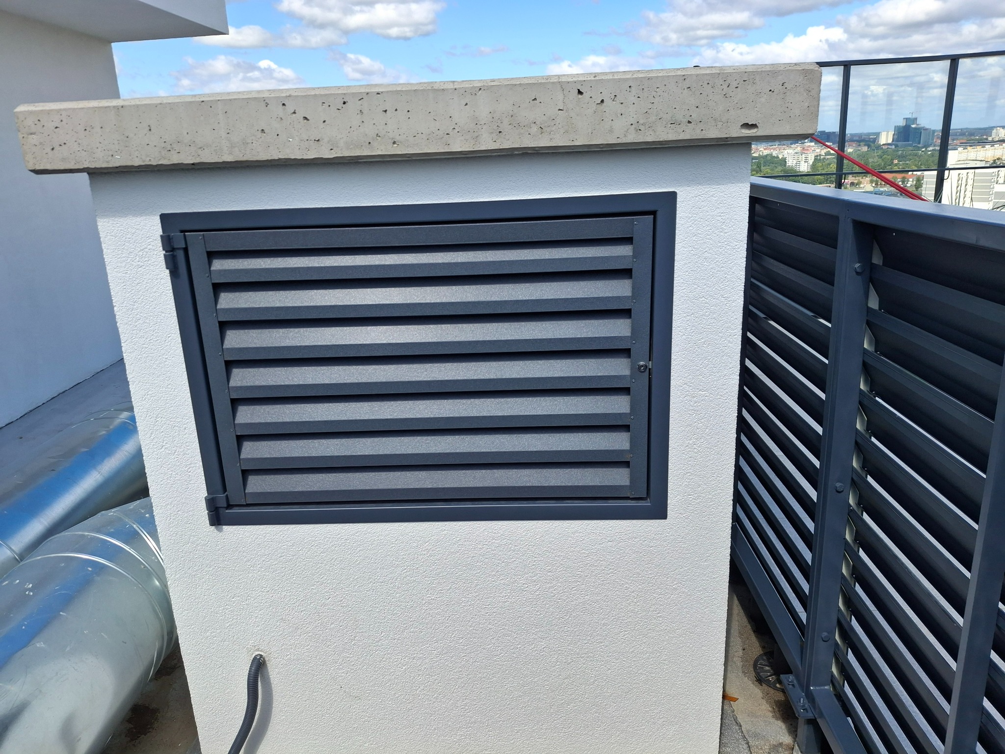 Szara metalowa żaluzja wentylacyjna na białej ścianie budynku, widok z zewnątrz na tle błękitnego nieba i zabudowy miejskiej.