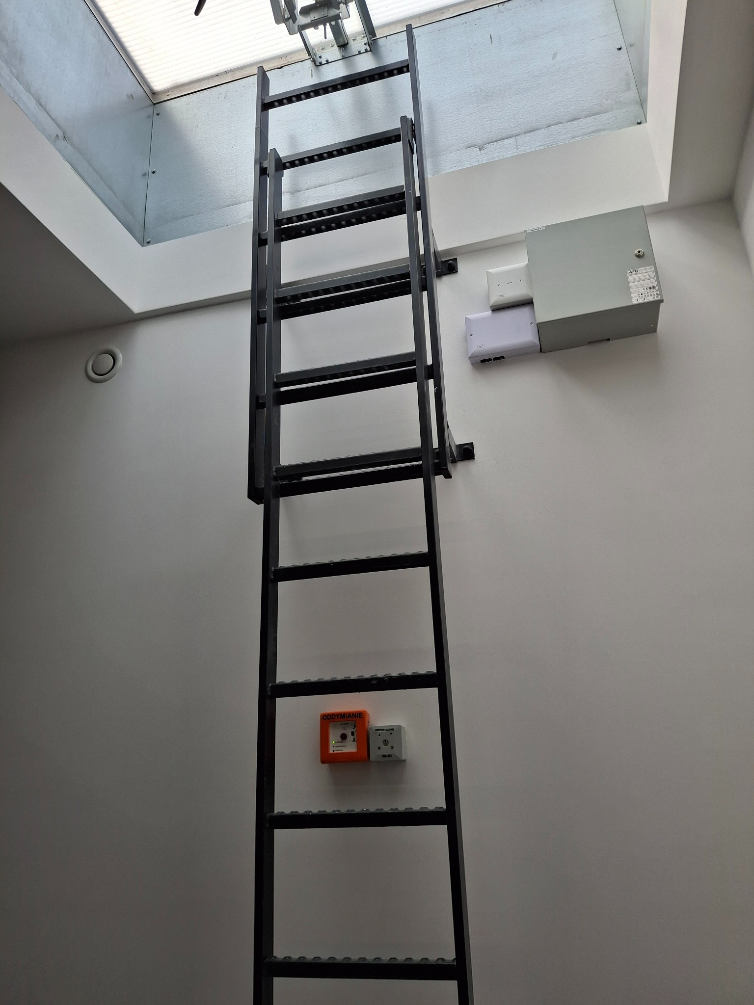 Metalowa drabina przymocowana do ściany prowadząca do wyłazu dachowego, widoczny system otwierania wyłazu, obok drabiny na ścianie zamontowane urządzenia elektryczne i czujnik oddymiania.