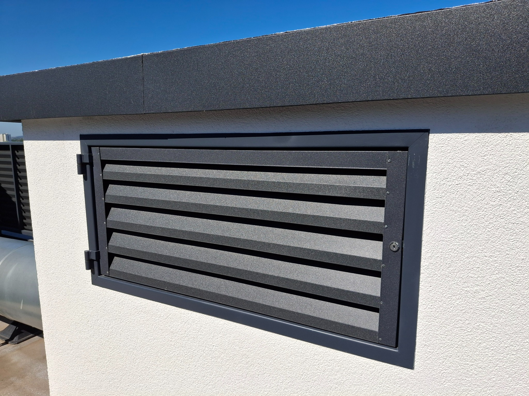 Szara metalowa żaluzja wentylacyjna zamykana na klucz, wkomponowana w elewację budynku, widoczna z bliska w słoneczny dzień.