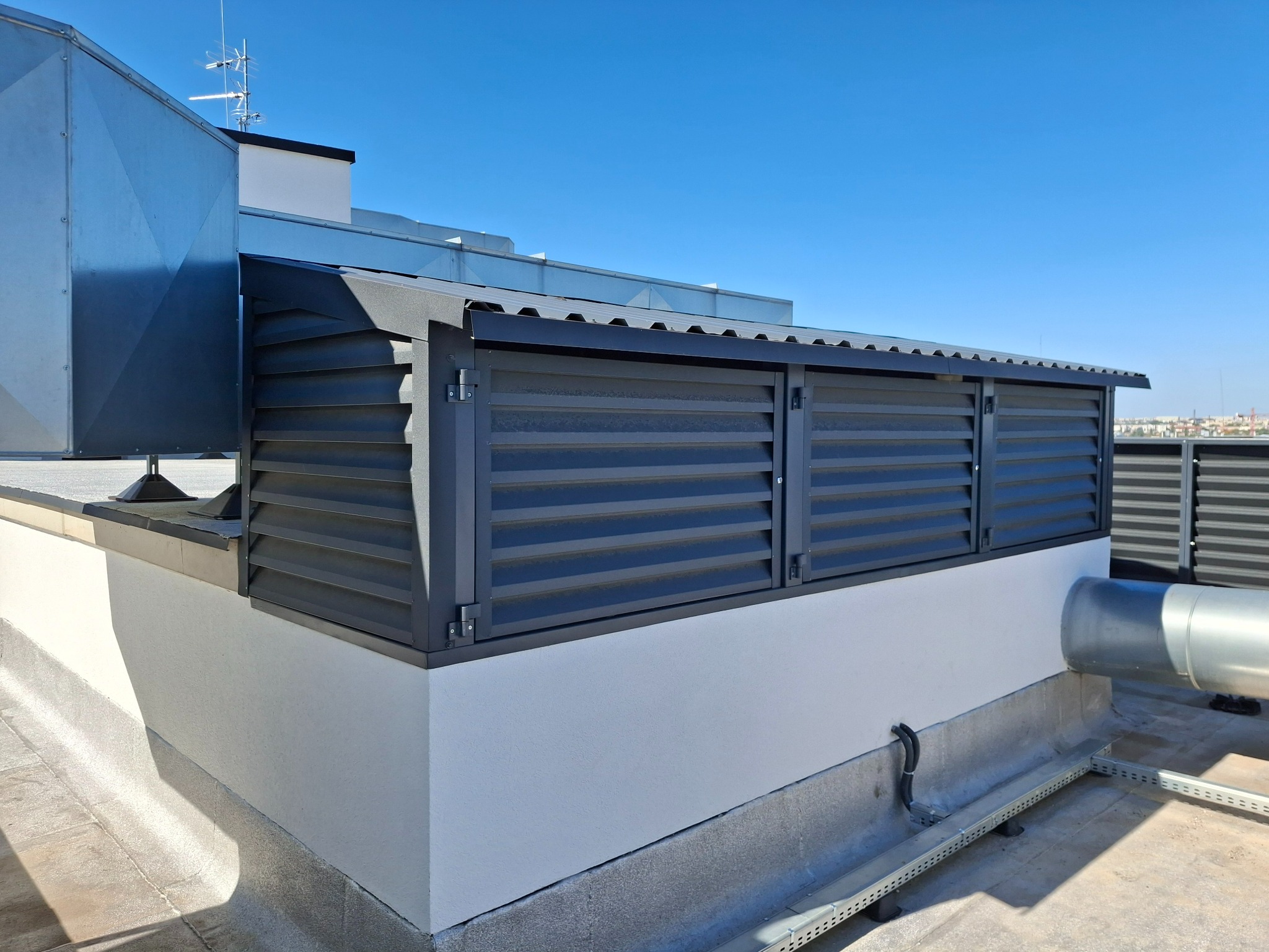 Szare, poziome panele osłonowe na dachu budynku, z widocznymi elementami wentylacji i anteną na tle błękitnego nieba.