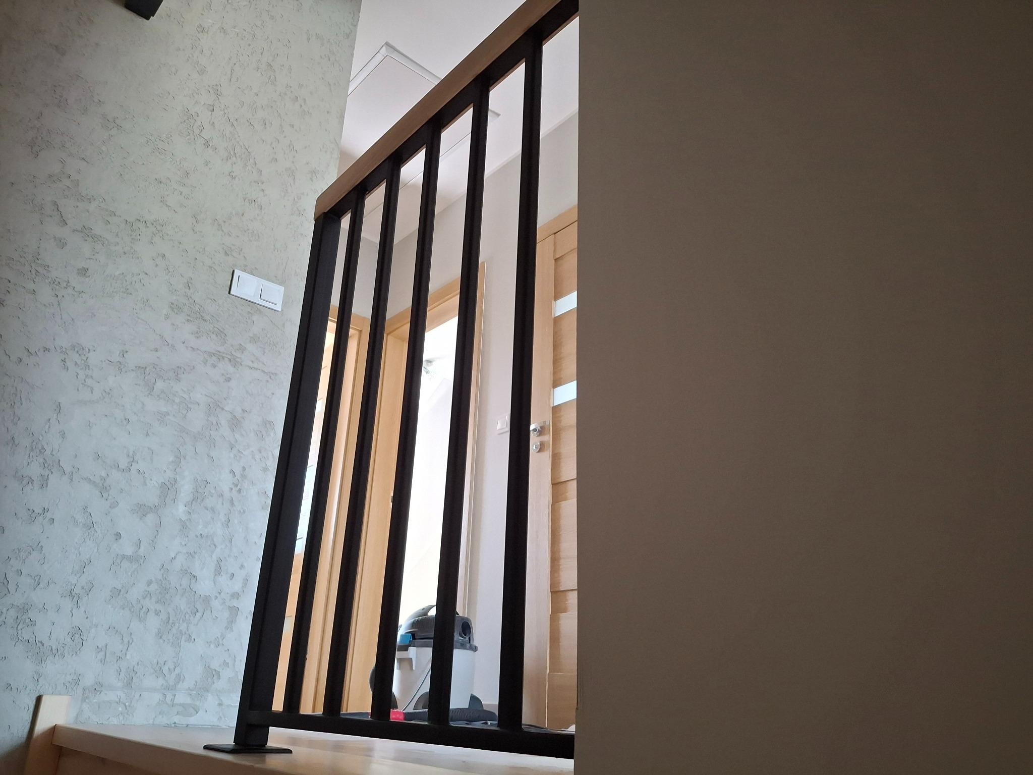 Nowoczesna balustrada schodowa z czarnymi, pionowymi prętami i drewnianym pochwytem, widok z dołu schodów na jasne drzwi i odkurzacz w tle.