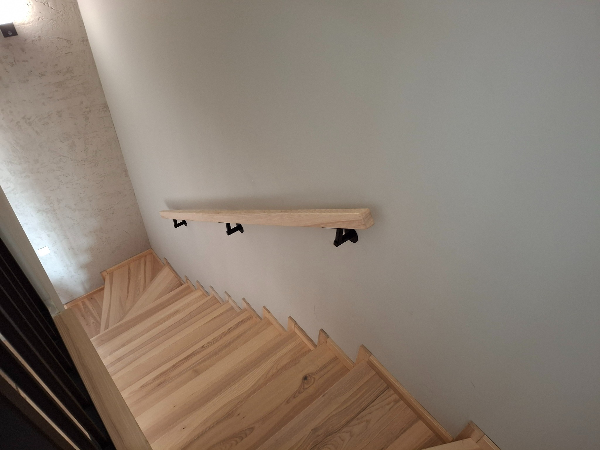 Drewniane schody z jasnego drewna z zamontowaną przyścienną, surową, drewnianą poręczą na czarnych wspornikach, widok z góry.