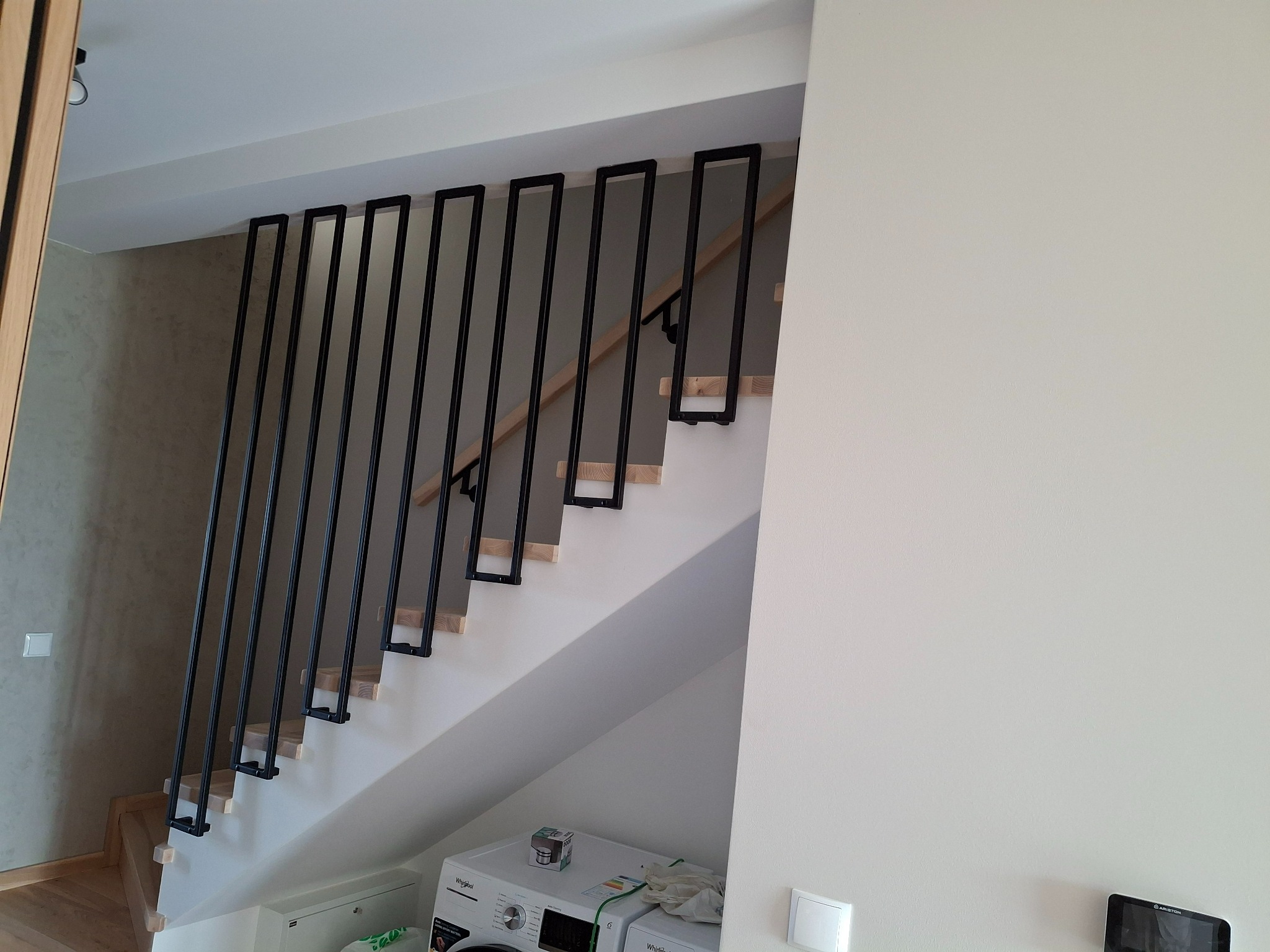 Nowoczesne schody z drewnianymi stopniami i czarną, minimalistyczną balustradą z prostokątnych elementów, widoczne z dołu, z pralką w tle.