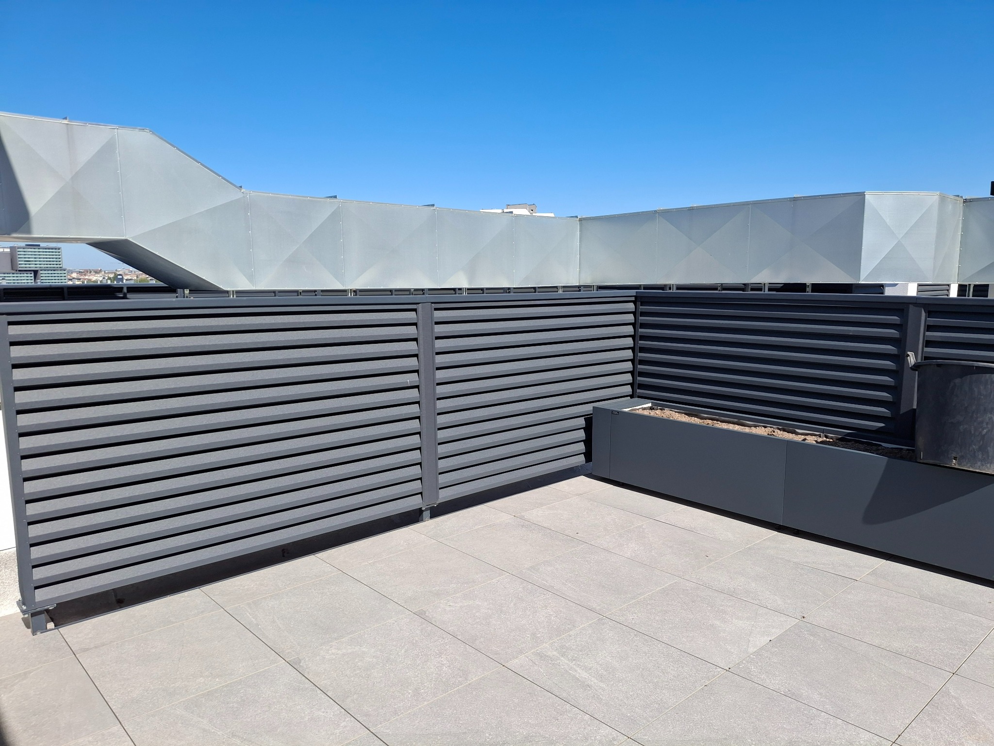 Szare poziome panele ogrodzeniowe na tle błękitnego nieba i zabudowań miejskich, z donicą na rośliny w rogu.