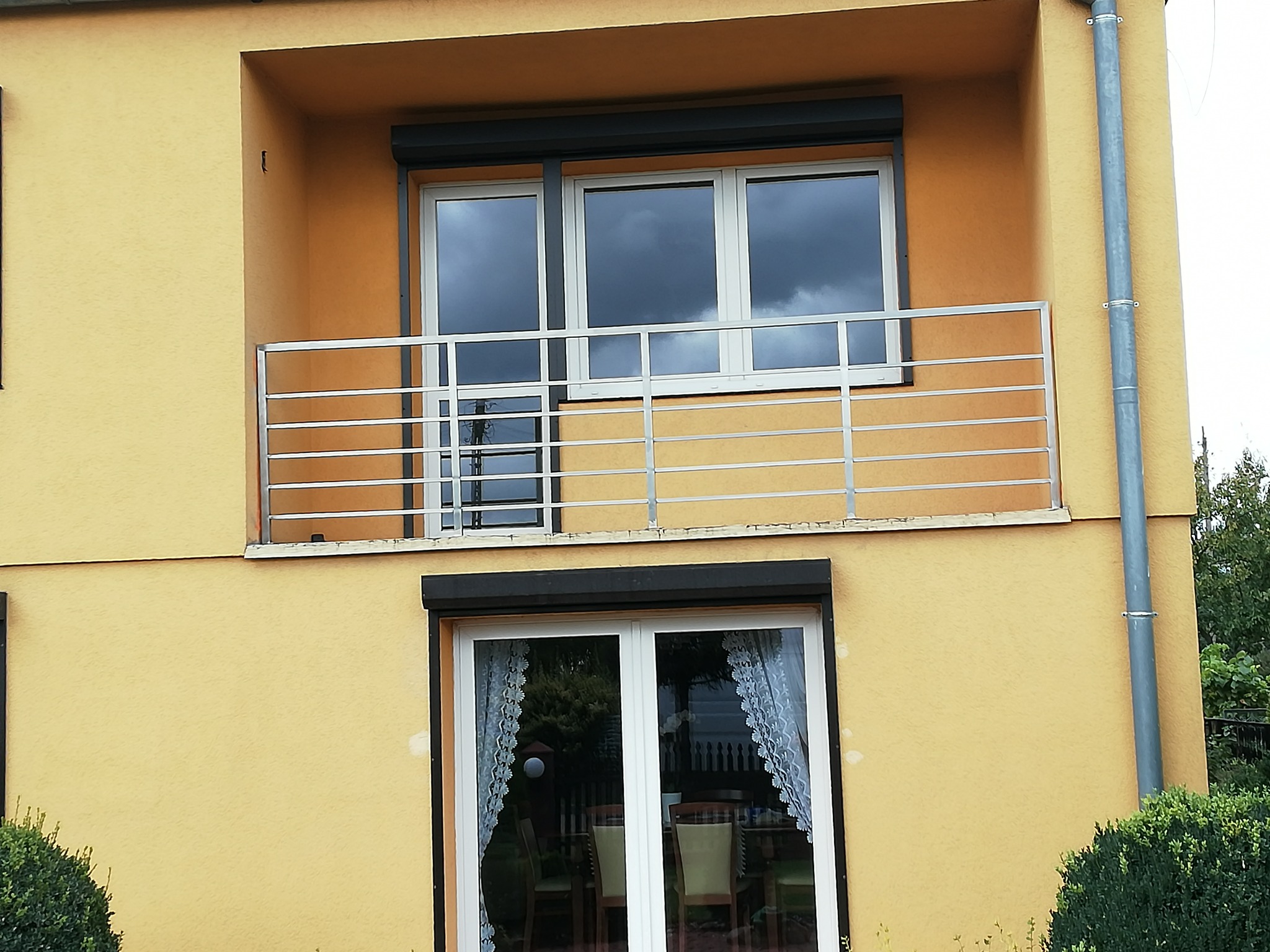 Żółta elewacja domu z balkonem zabezpieczonym prostą, poziomą, stalową konstrukcją, okna z roletami i firankami.