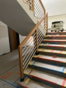 Montaż drewnianej poręczy z metalowymi tralkami na schodach z widocznymi śladami prac wykończeniowych i odkurzaczem przemysłowym w tle.