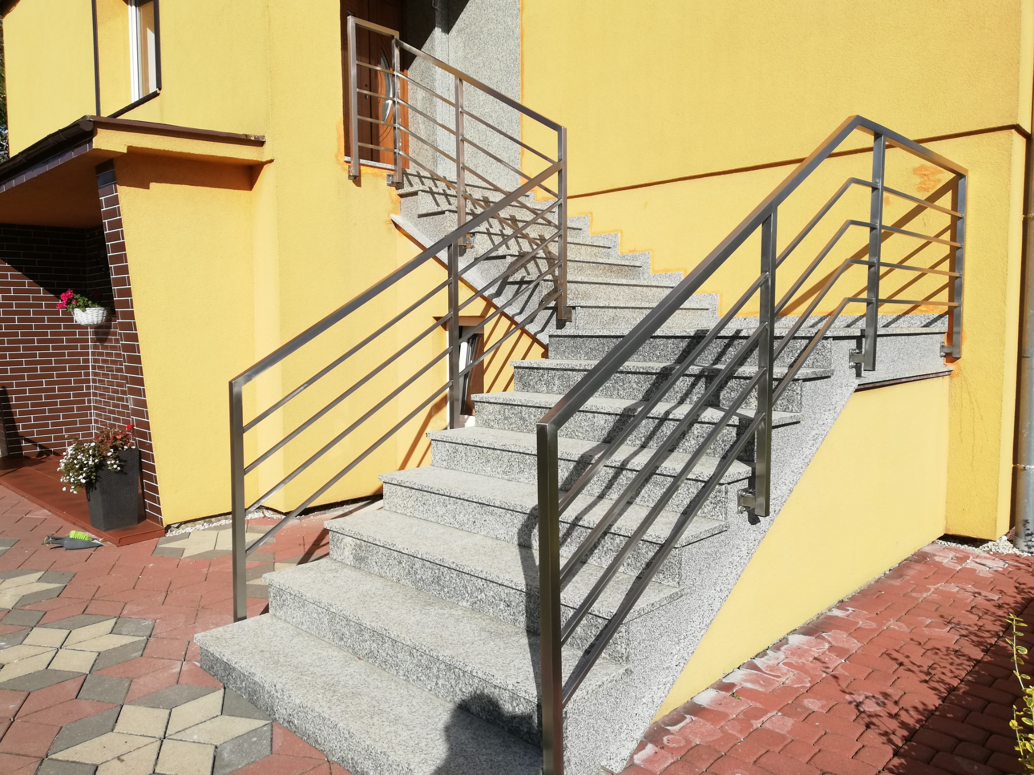 Zewnętrzne schody z szarego granitu prowadzące do drzwi wejściowych, zabezpieczone prostą, nowoczesną balustradą ze stali nierdzewnej, na tle żółtej elewacji budynku.