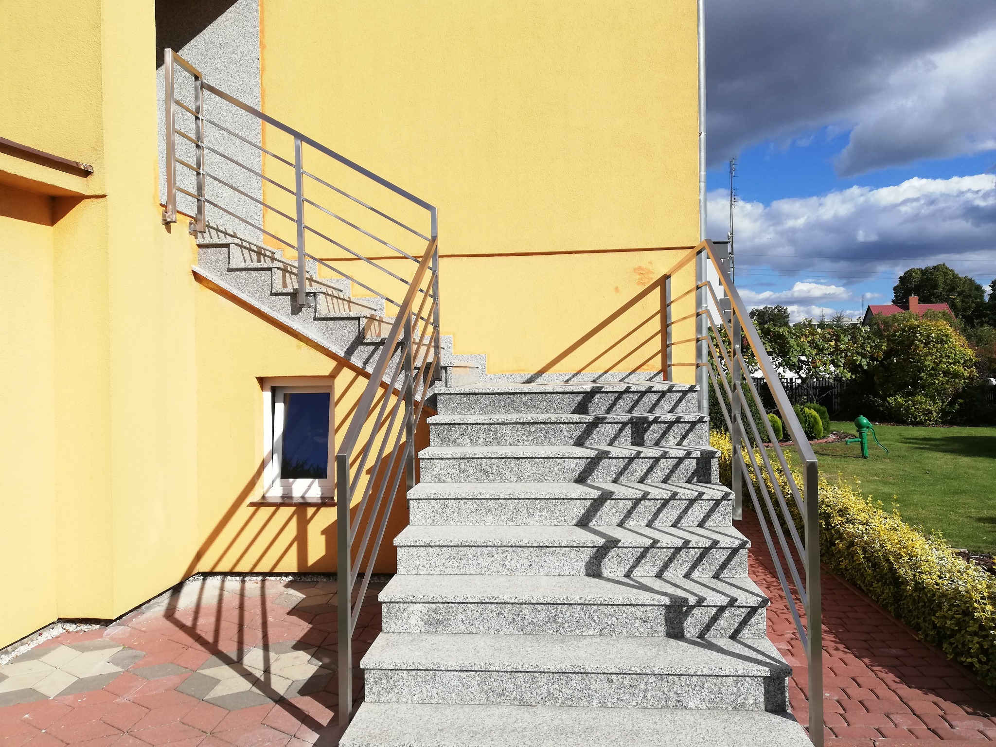 Zewnętrzne schody z szarego granitu z nierdzewną balustradą, prowadzące na piętro budynku o żółtej elewacji, z widocznym oknem i fragmentem ogrodu w tle.
