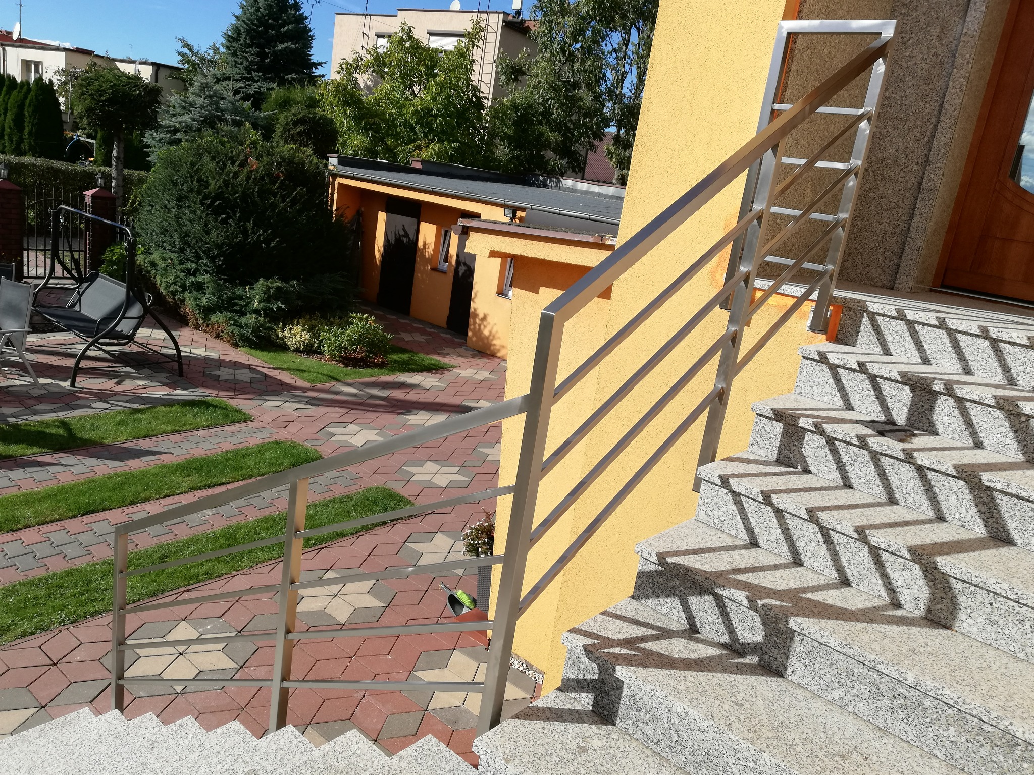 Zewnętrzne schody granitowe z balustradą ze stali nierdzewnej, prowadzące do wejścia budynku o żółtej elewacji, widok z góry na ozdobny chodnik z kostki i trawy w tle.