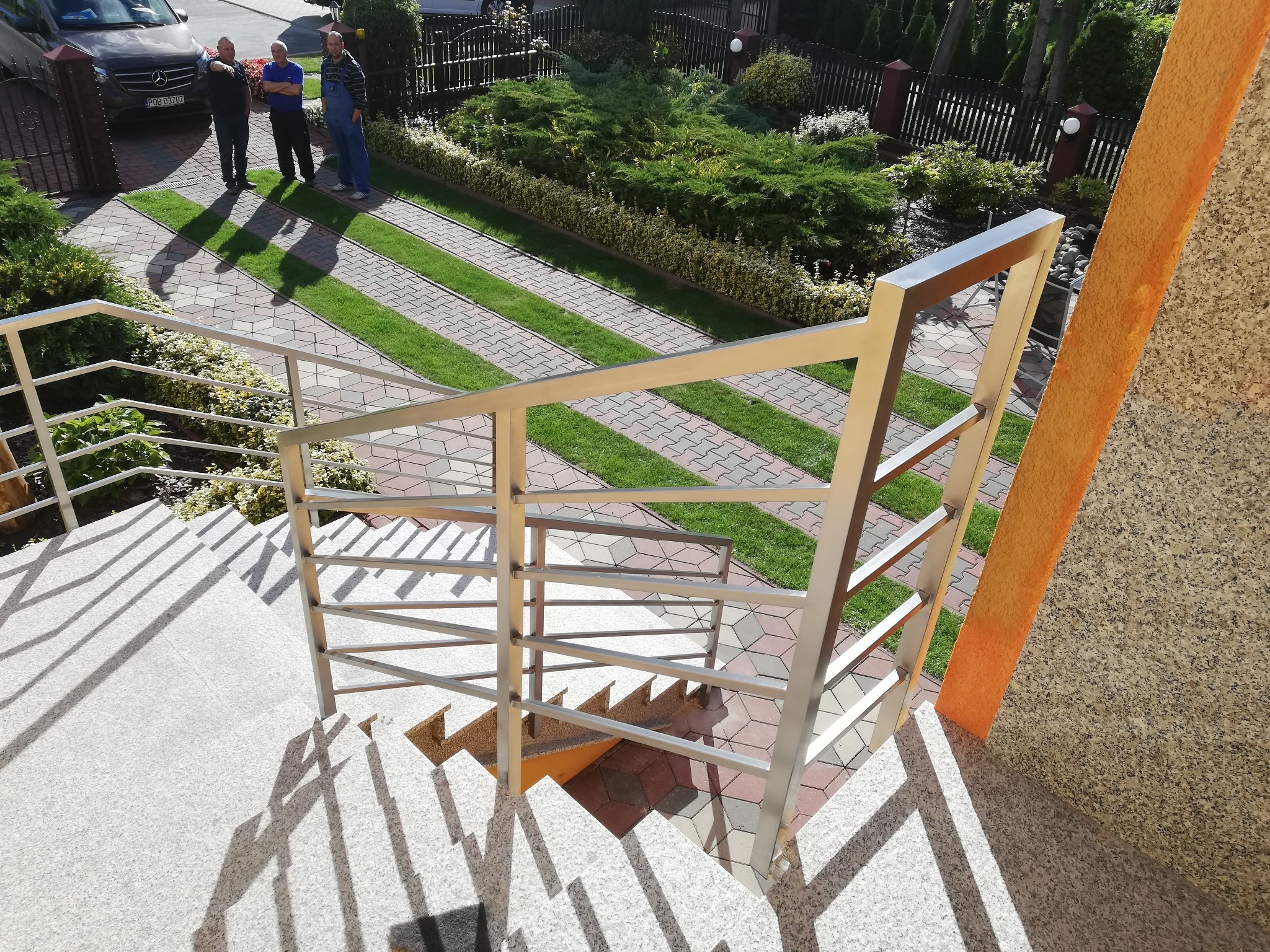 Zewnętrzne schody z jasnego kamienia z nowoczesną, prostą balustradą ze stali nierdzewnej, widok z góry na podjazd z kostki i trawy oraz grupę osób przed domem.