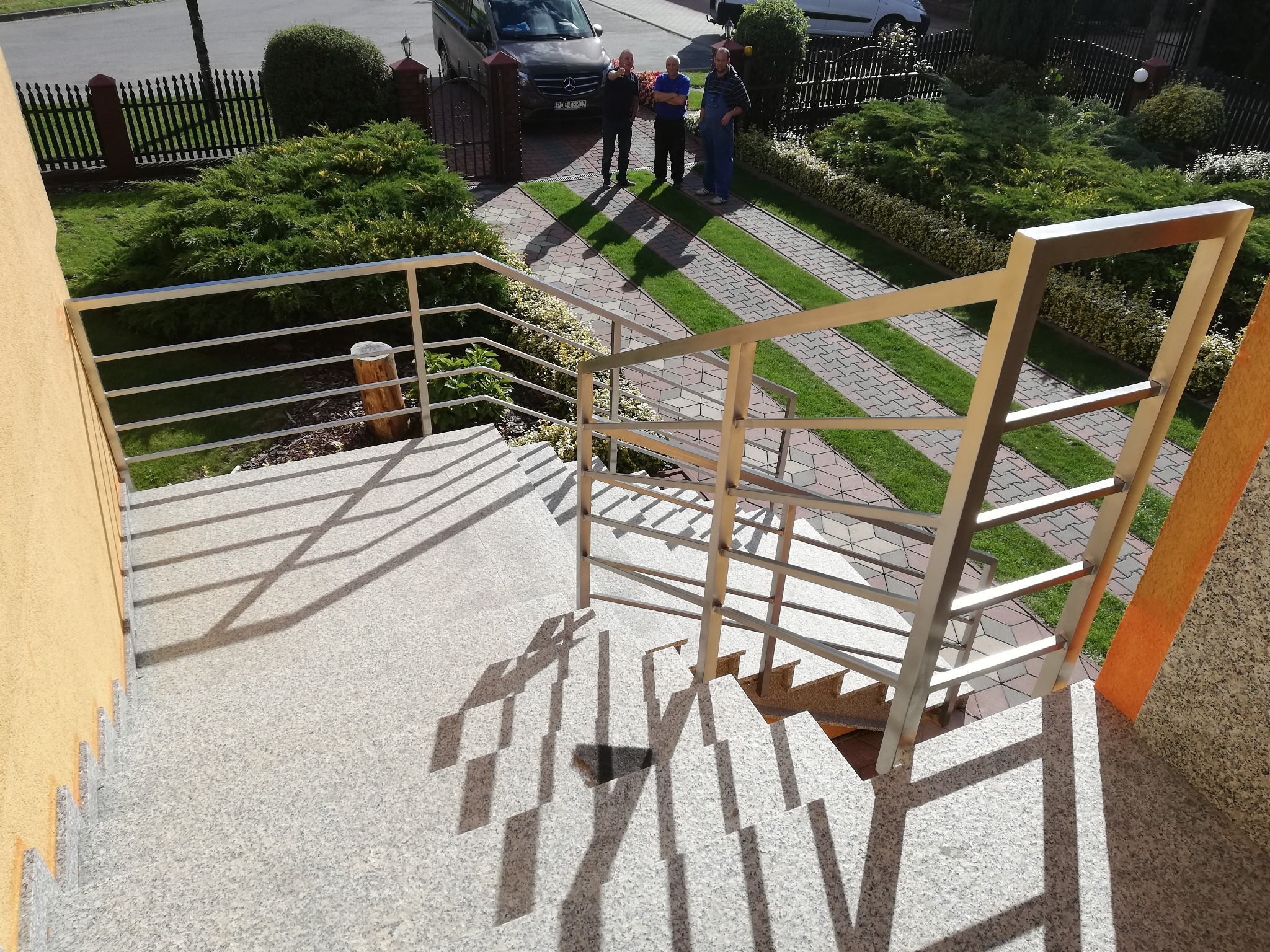 Zewnętrzne schody z jasnego granitu z nowoczesną balustradą ze stali nierdzewnej, widok z góry na ogród i trzech mężczyzn przed domem.