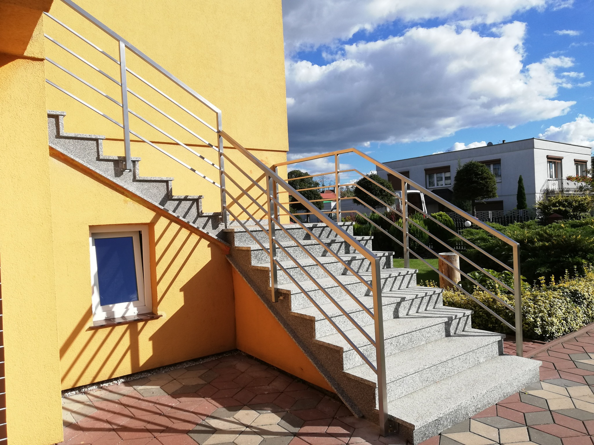Zewnętrzne schody z szarego granitu z nierdzewną balustradą, prowadzące do wejścia budynku o żółtej elewacji, widoczne w słoneczny dzień na tle błękitnego nieba z chmurami.