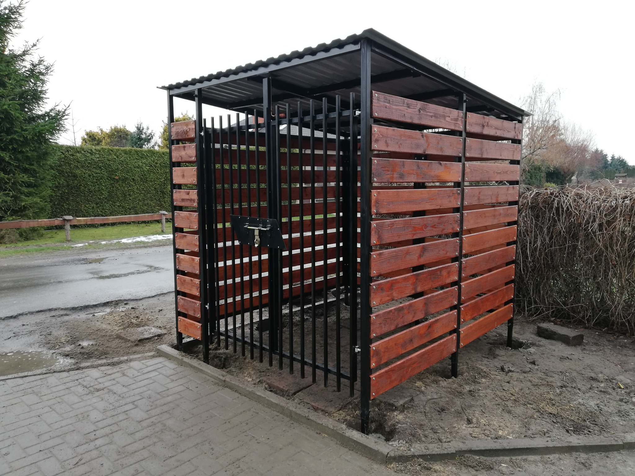 Zadaszona wiata śmietnikowa z metalową konstrukcją i drewnianymi panelami w kolorze brązowym, zamykana na zamek, na tle ogrodu w pochmurny dzień.