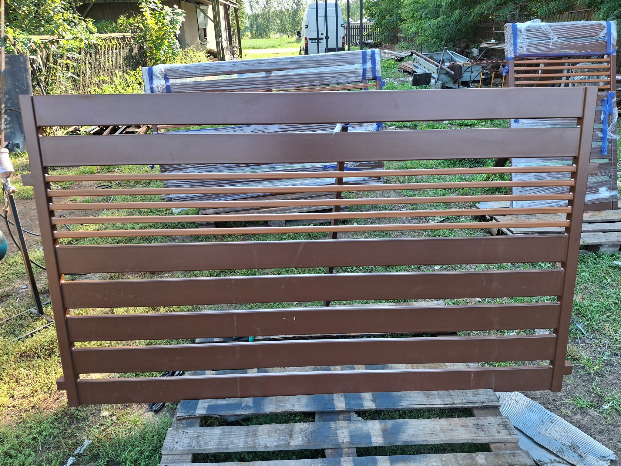 Gotowy, brązowy panel ogrodzeniowy z poziomymi szczeblami, leżący na palecie. W tle widoczne inne elementy ogrodzenia, częściowo owinięte folią.