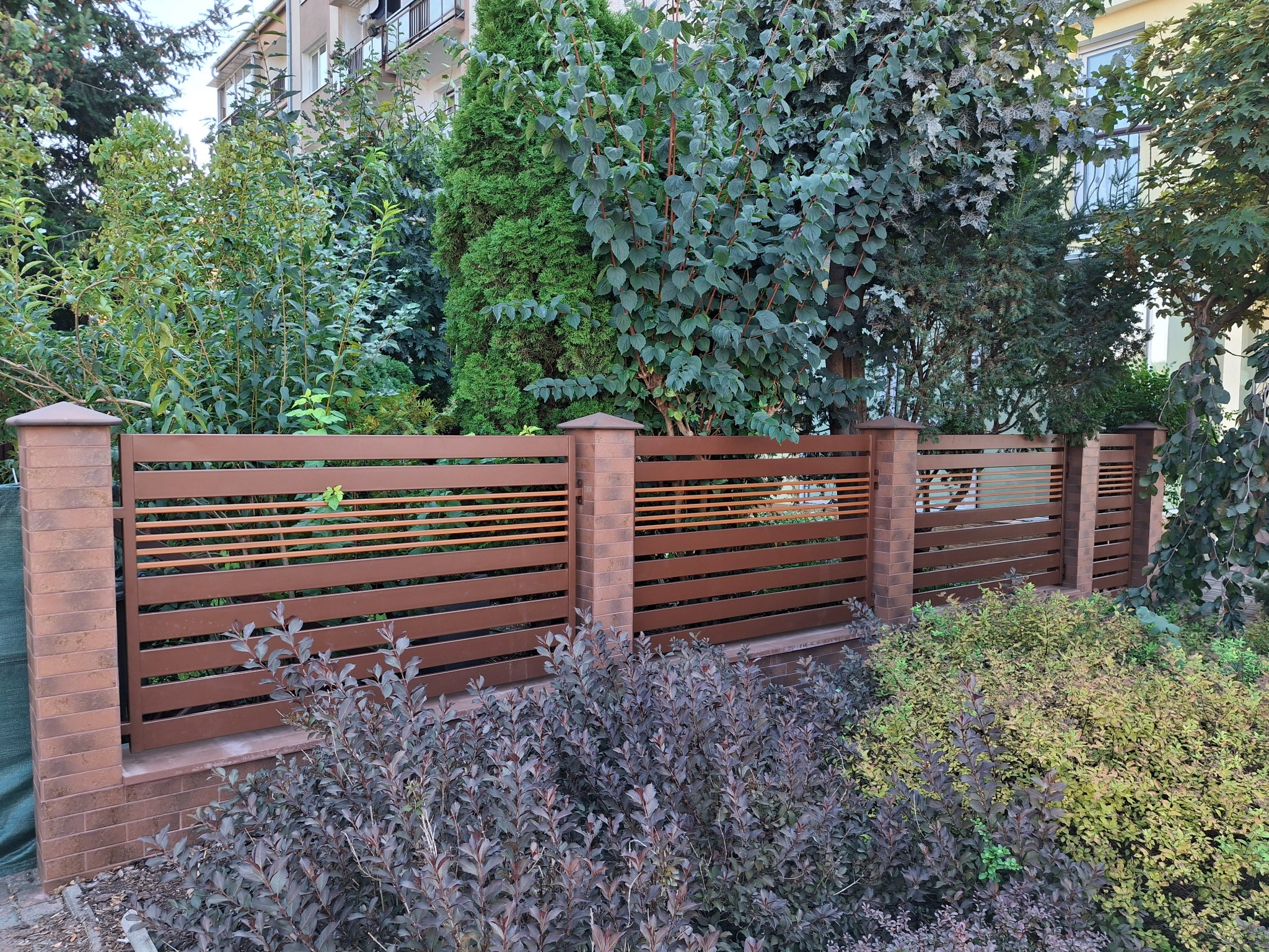 Nowoczesne ogrodzenie z poziomych, brązowych paneli i ceglanych słupków, otoczone zielenią. Widok na budynek w tle. Ujęcie z poziomu gruntu.