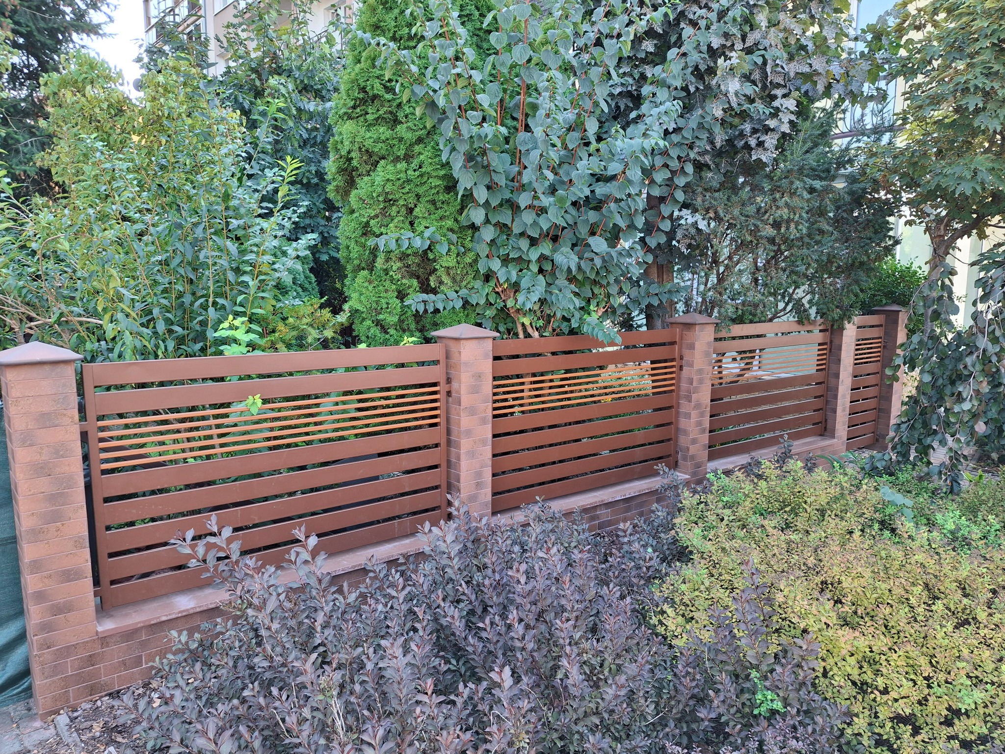 Nowoczesne ogrodzenie z poziomych, brązowych paneli metalowych, osadzone na ceglanych słupkach, otoczone zielenią. Widok z góry.