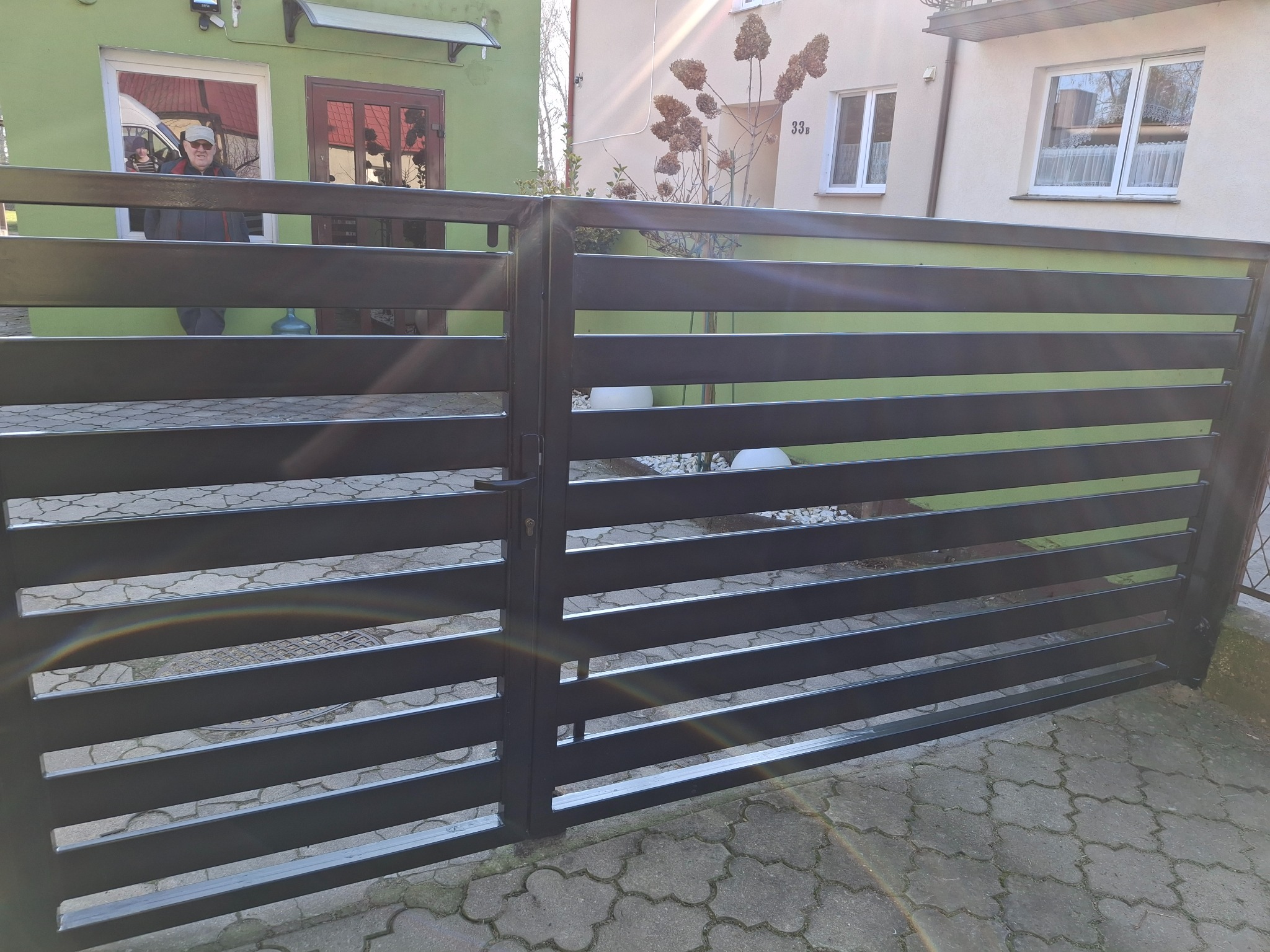 Nowoczesna, pozioma brama wjazdowa w kolorze antracytowym, z widocznym mechanizmem zamka. Tło stanowi budynek mieszkalny z zieloną elewacją i elementami dekoracyjnymi.