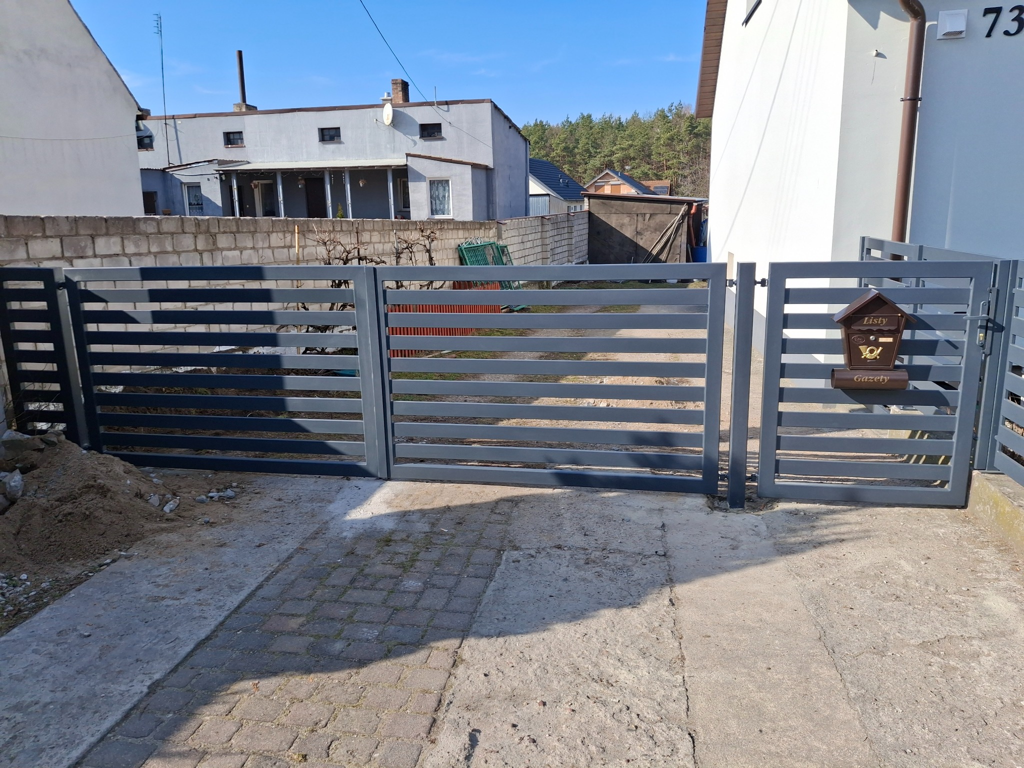 Nowoczesna, pozioma brama wjazdowa w kolorze szarym, z przylegającą furtką i brązową skrzynką na listy. W tle budynki mieszkalne i drzewa. Słoneczny dzień.