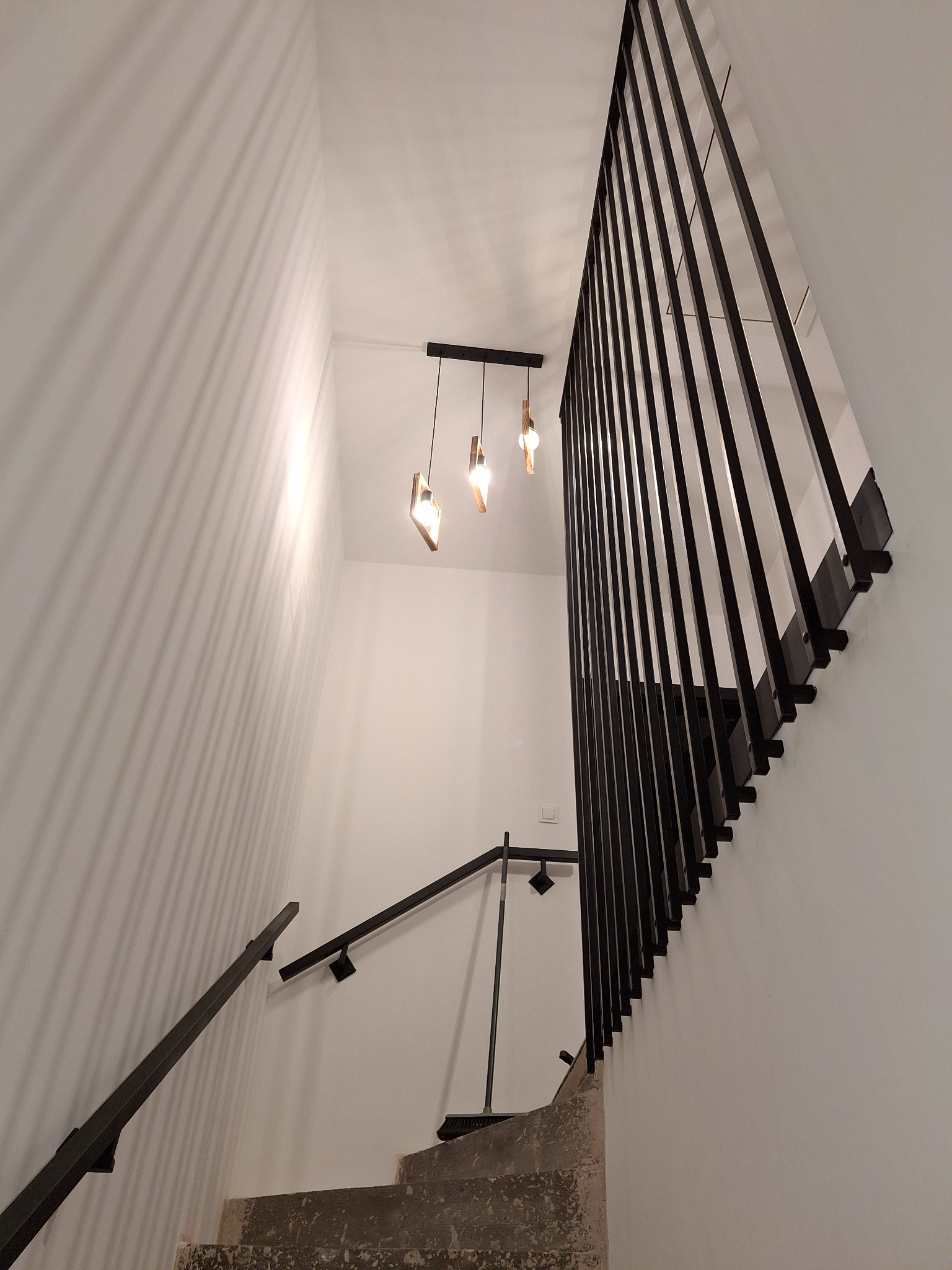 Nowoczesne schody z czarną, metalową balustradą w minimalistycznym wnętrzu. Uwagę przyciągają geometryczne lampy wiszące i gra światła na białych ścianach.