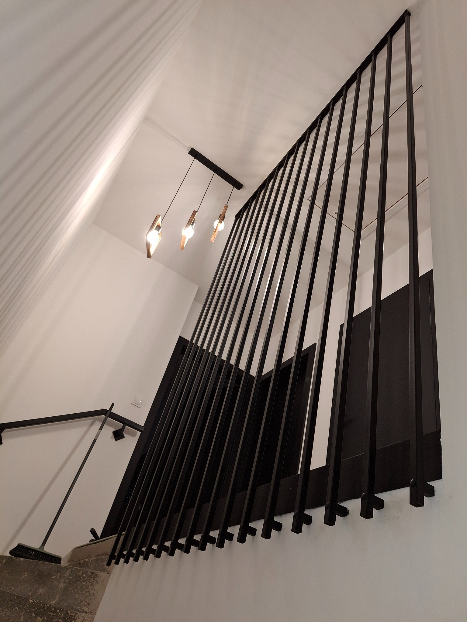 Minimalistyczna, czarna balustrada schodowa z pionowych prętów, oświetlona wiszącymi lampami. Ujęcie z dołu, podkreślające geometrię i nowoczesny design.