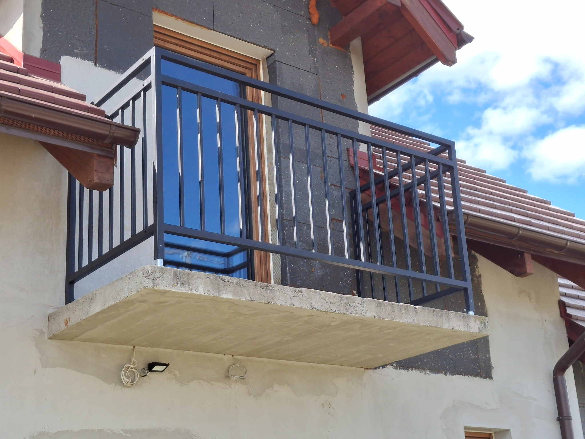 Czarna, metalowa balustrada balkonowa z pionowymi szczebelkami, zamontowana na betonowej płycie balkonu. Widok z dołu, fragment elewacji budynku w tle.