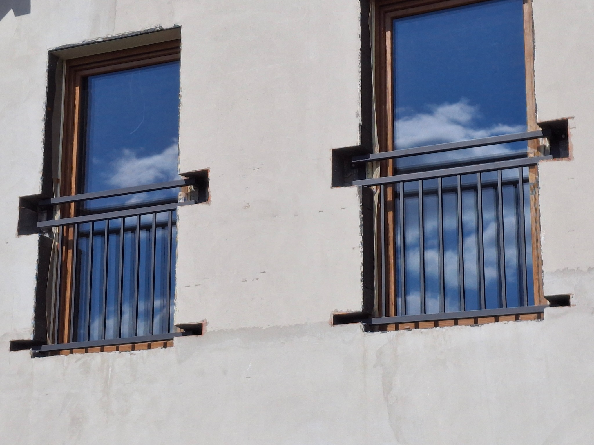 Dwa okna z zamontowanymi, nowoczesnymi, metalowymi balustradami balkonowymi w kolorze grafitowym, w trakcie montażu na surowej elewacji budynku. Widoczne otwory montażowe.