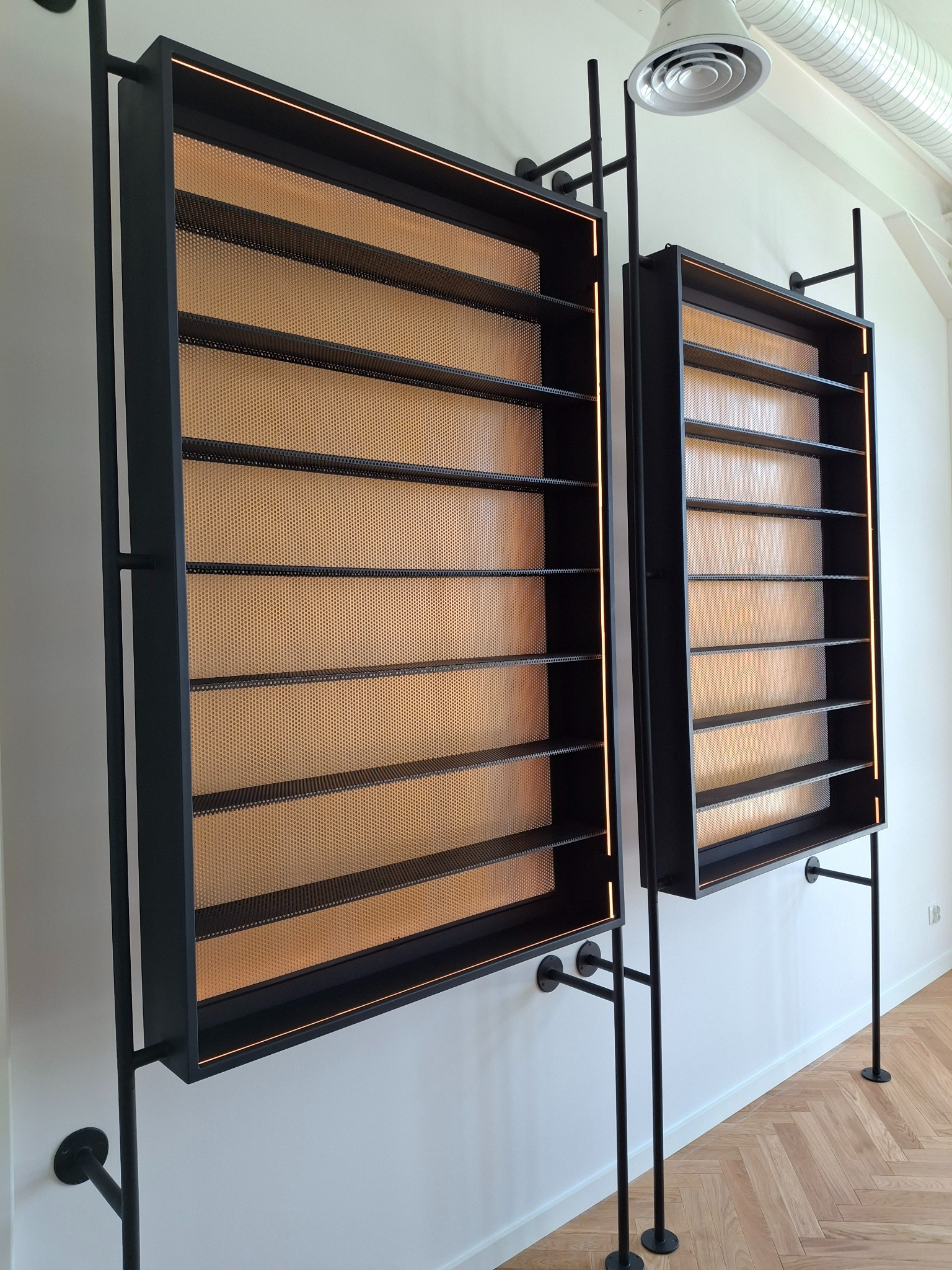 Dwie metalowe, czarne półki z perforowanym złotym tłem, zamontowane na ścianie. Minimalistyczny design z oświetleniem LED. Widoczny fragment podłogi z parkietem.