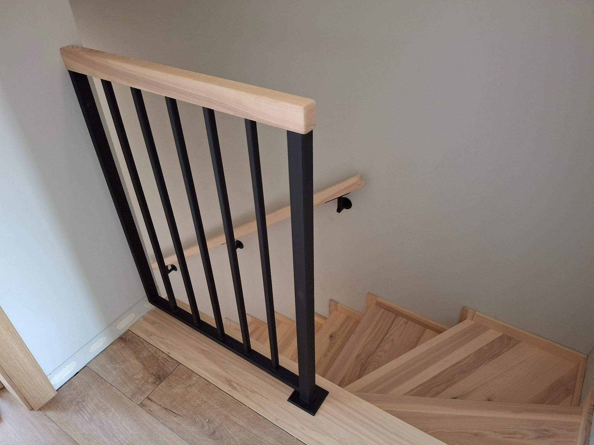 Nowoczesna balustrada schodowa z czarnymi, metalowymi słupkami i jasnym, drewnianym poręczą, widziana z góry, podkreślająca minimalistyczny design i naturalne materiały.