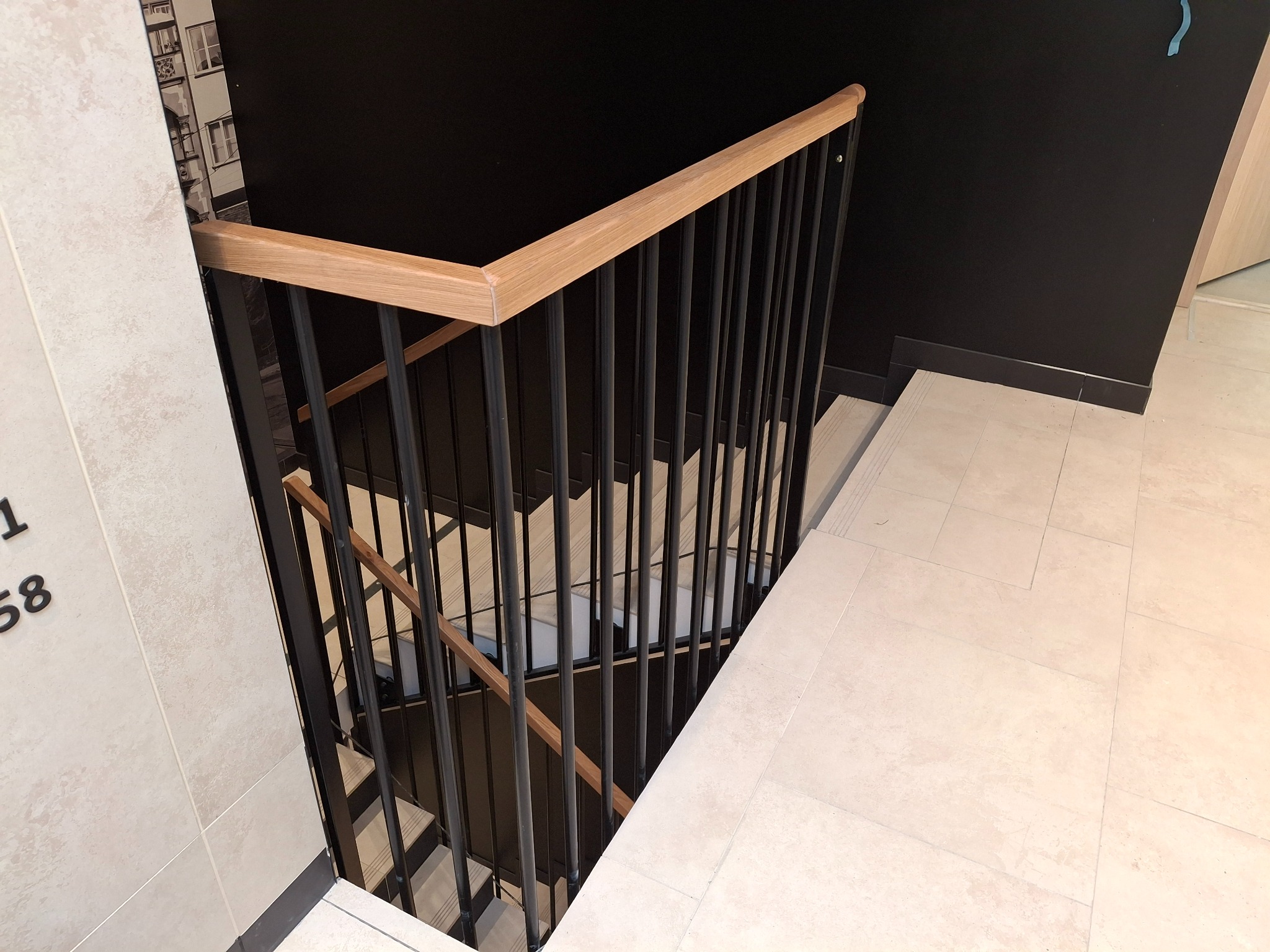 Nowoczesne schody z czarną metalową balustradą i drewnianym poręczą, widok z góry na jasne stopnie i ciemną ścianę. Minimalistyczny design.