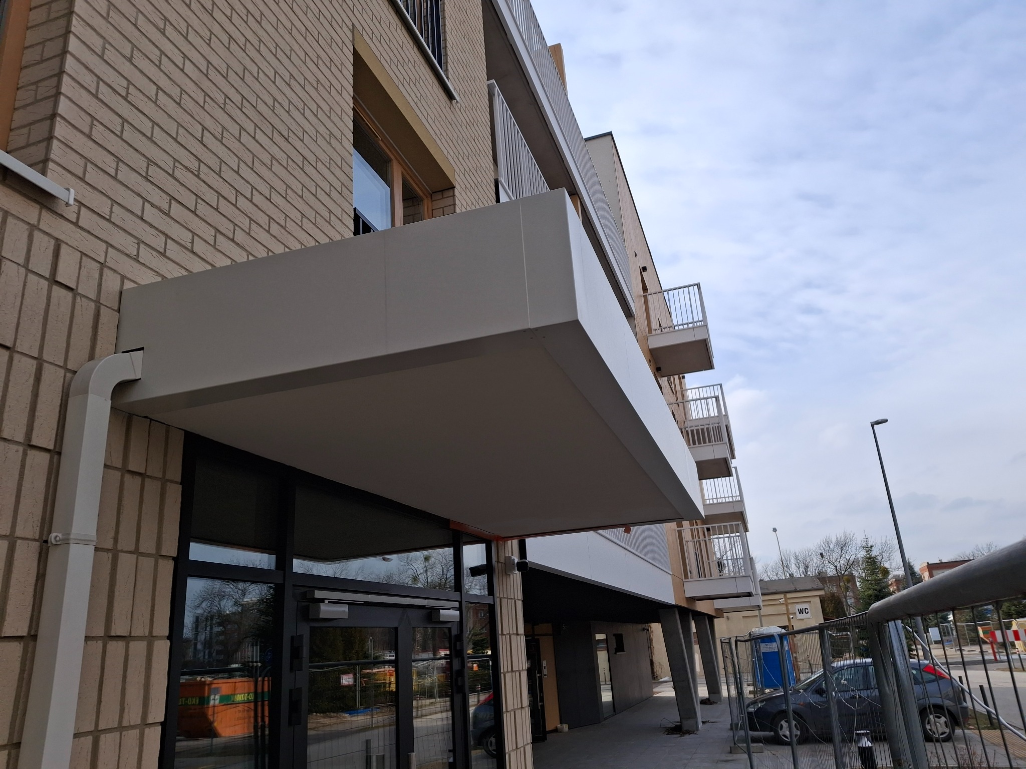 Nowoczesny budynek z balkonami o minimalistycznej architekturze. Jasna elewacja, metalowe balustrady i duże okna tworzą spójną całość. Widoczne detale wykończeniowe.