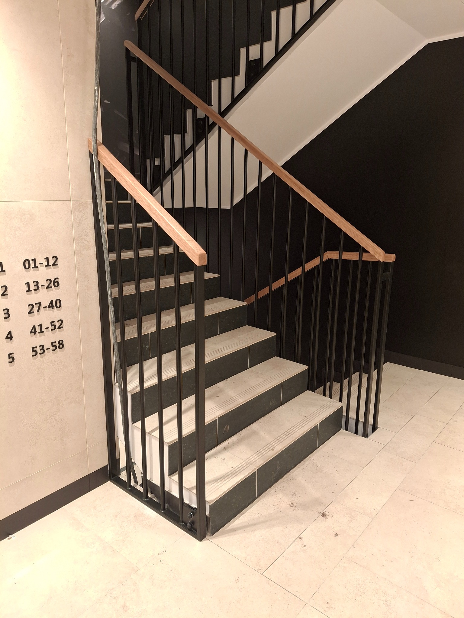 Metalowe schody z drewnianą poręczą w nowoczesnym budynku. Widoczne numery mieszkań na ścianie. Minimalistyczny design, czarne barierki i jasne stopnie.