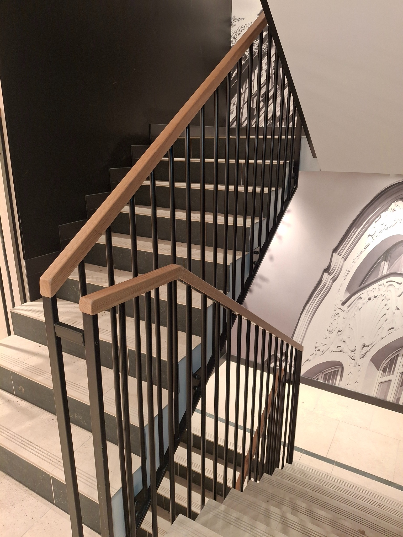 Nowoczesne schody z czarną metalową balustradą i drewnianym poręczą, widok z góry. Na ścianie dekoracyjny motyw architektoniczny w odcieniach szarości.