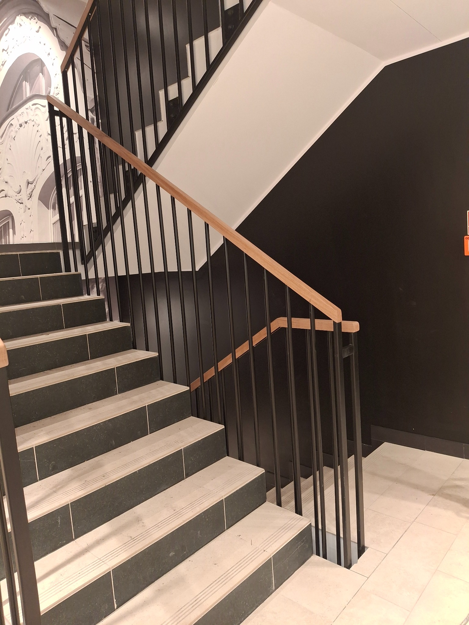 Nowoczesne schody z czarną metalową balustradą i drewnianym poręczą, na tle czarnej ściany i dekoracyjnej grafiki. Widok z dołu, podkreślający minimalistyczny design.