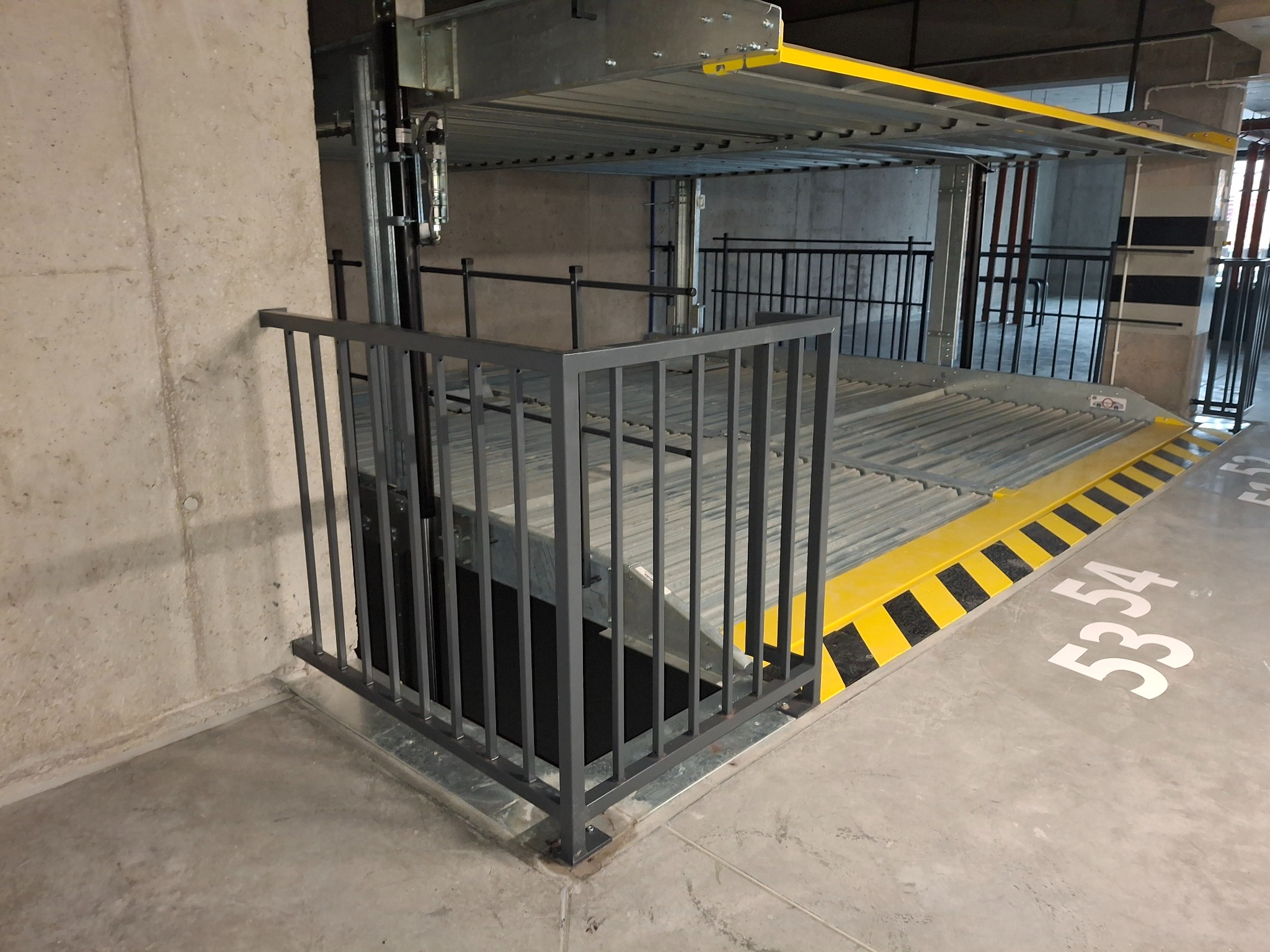 Szara, metalowa barierka zabezpieczająca platformę parkingu wielopoziomowego. Żółto-czarne pasy ostrzegawcze na krawędzi platformy. Numeracja miejsc parkingowych na posadzce.